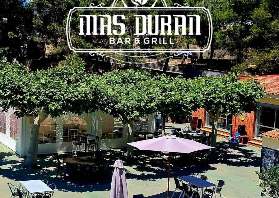 Terrassa exterior del Bar&Grill Mas Duran, amb taules sota els arbres i para-sols, i el logotip de l'establiment sobreimprès a la part superior