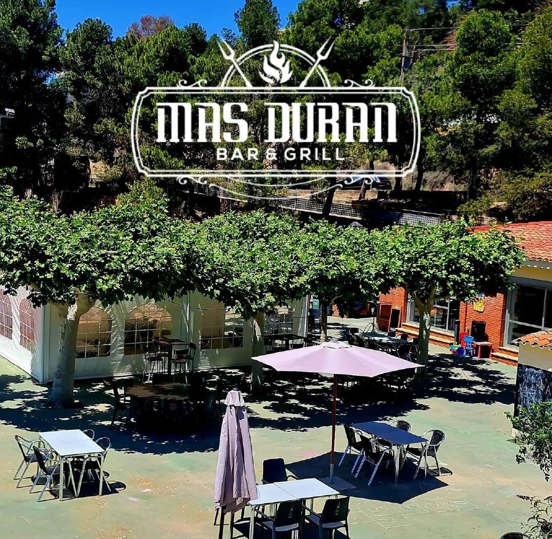 Terrassa exterior del Bar&Grill Mas Duran, amb taules sota els arbres i para-sols, i el logotip de l'establiment sobreimprès a la part superior