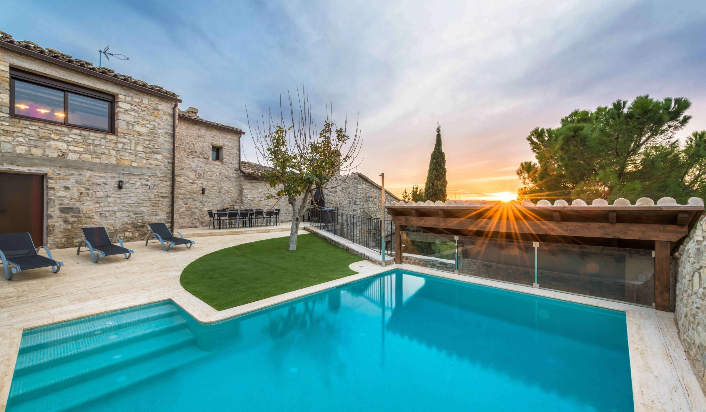 Piscina exterior de Cal Rovira amb gandules i gespa, al costat de la casa de pedra i una pèrgola de fusta, amb la posta de sol al fons