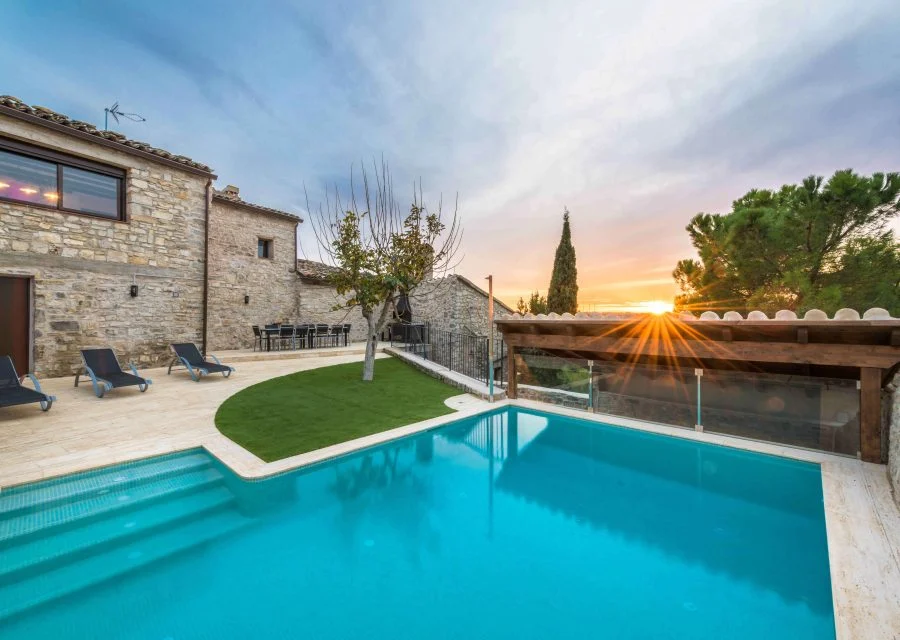 Piscina exterior de Cal Rovira amb gandules i gespa, al costat de la casa de pedra i una pèrgola de fusta, amb la posta de sol al fons