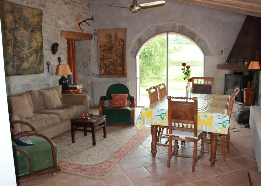 Sala d'estar i menjador de Cal Tonillo i Cal Tacies, amb murs de pedra vista, un tapís, sofàs, taula parada i un finestral d'arc que dona al camp