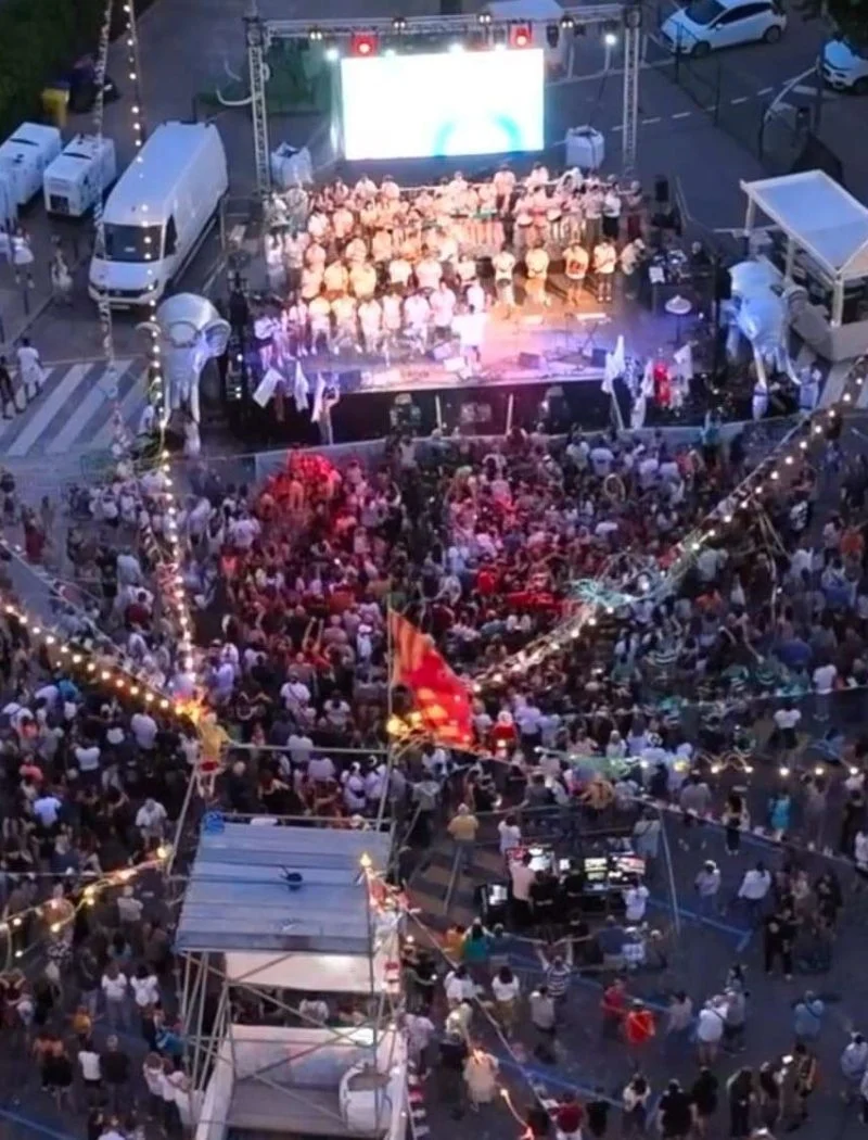 Vista aèria nocturna del Carnaval de Secà, amb una comparsa disfressada damunt l'escenari il·luminat i una gran gentada omplint el carrer sota les garlandes de bombetes