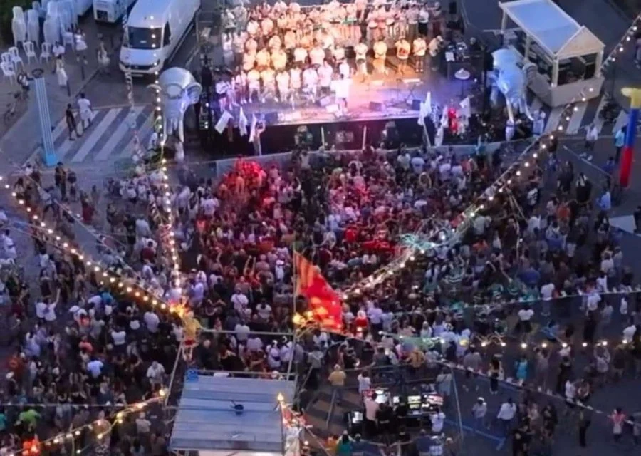 Vista aèria nocturna del Carnaval de Secà, amb una comparsa disfressada damunt l'escenari il·luminat i una gran gentada omplint el carrer sota les garlandes de bombetes
