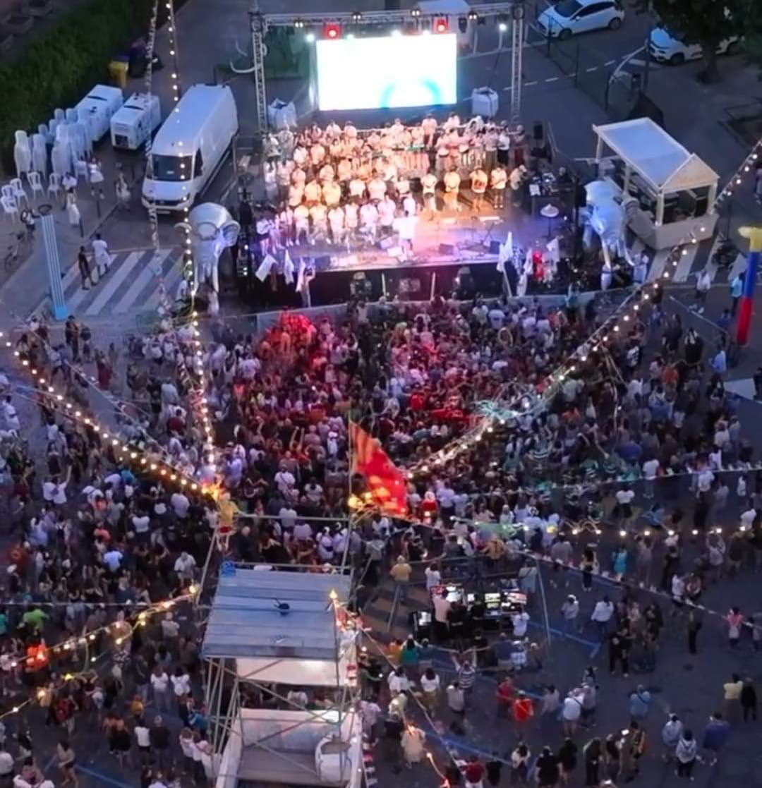Vista aèria nocturna del Carnaval de Secà, amb una comparsa disfressada damunt l'escenari il·luminat i una gran gentada omplint el carrer sota les garlandes de bombetes