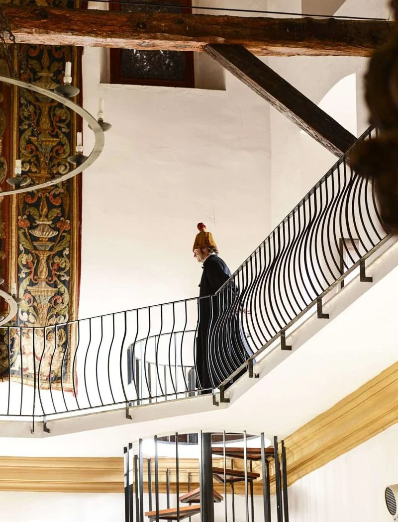 Visita teatralitzada a la Universitat de Cervera: una persona recorre la barana d'una planta noble amb bigues de fusta vistes, al costat d'un gran tapís i una escala de cargol