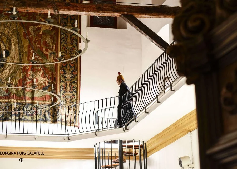 Visita teatralitzada a la Universitat de Cervera: una persona recorre la barana d'una planta noble amb bigues de fusta vistes, al costat d'un gran tapís i una escala de cargol