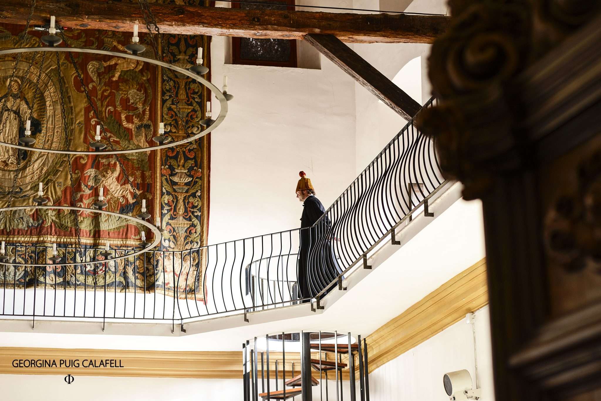 Visita teatralitzada a la Universitat de Cervera: una persona recorre la barana d'una planta noble amb bigues de fusta vistes, al costat d'un gran tapís i una escala de cargol