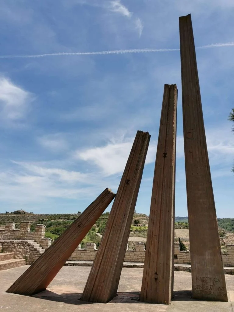 Monument a la Generalitat, de Josep M. Subirachs: quatre pilars metàl·lics inclinats de diferents alçades sobre una plataforma pavimentada, amb el paisatge de la Segarra al fons