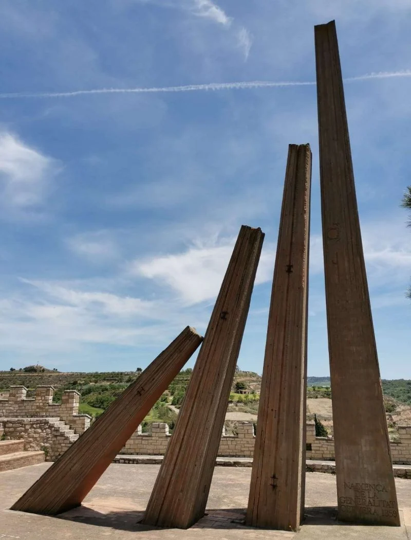 Monument a la Generalitat, de Josep M. Subirachs: quatre pilars metàl·lics inclinats de diferents alçades sobre una plataforma pavimentada, amb el paisatge de la Segarra al fons