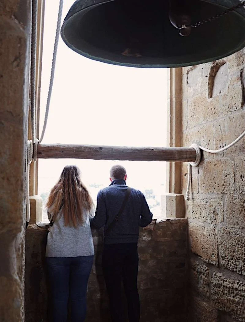 Dues persones d'esquena s'aboquen a la finestra del campanar de Santa Maria de Cervera per contemplar la panoràmica, amb una campana de bronze penjada al damunt