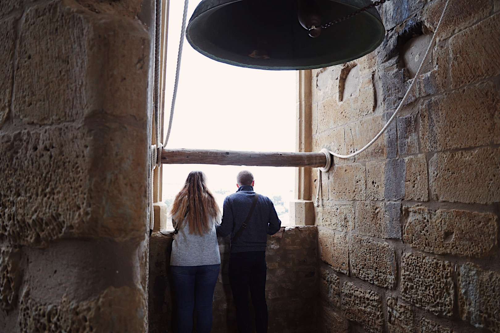 Dues persones d'esquena s'aboquen a la finestra del campanar de Santa Maria de Cervera per contemplar la panoràmica, amb una campana de bronze penjada al damunt