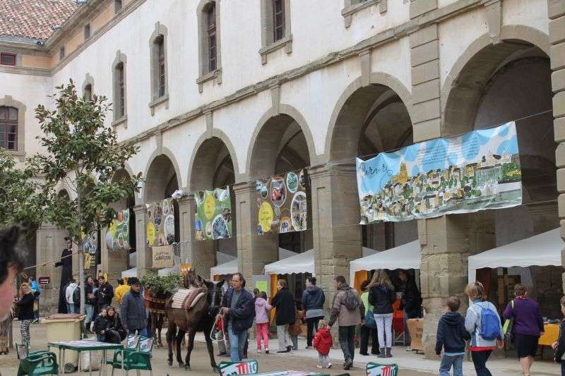 Parades de la Fira del Pa i el Cereal sota els arcs del pati de la Universitat de Cervera, amb visitants, una mula guarnida i pancartes il·lustrades penjades entre les arcades