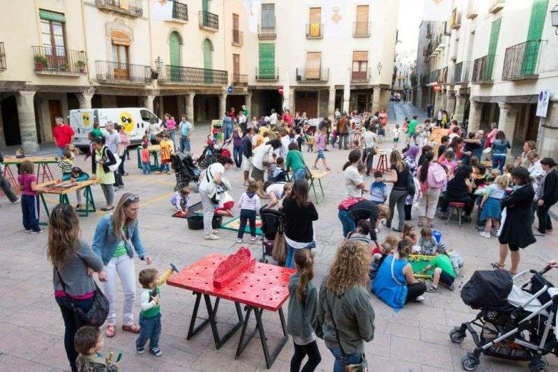 Activitats infantils de la Isagoge a la plaça porxada de Cervera, amb famílies, tallers en taules de fusta i canalla jugant entre els edificis de balcons