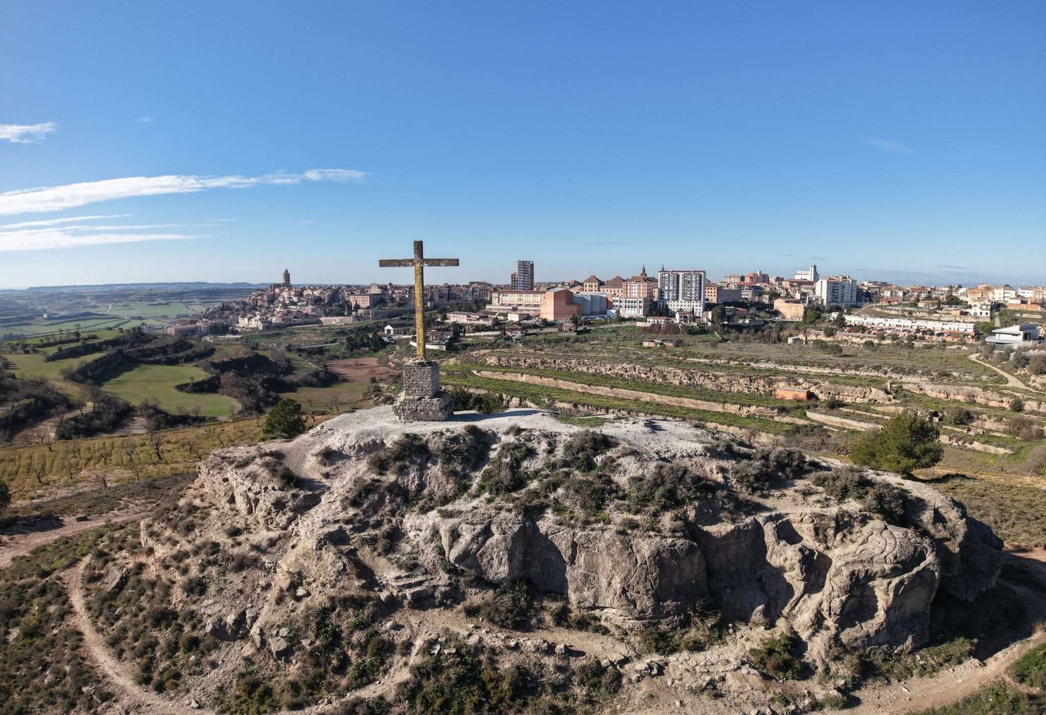 Vista aèria del turó rocós de les Forques, coronat per una gran creu de fusta, amb els camps de la Segarra a l'esquerra i el nucli de Cervera al fons