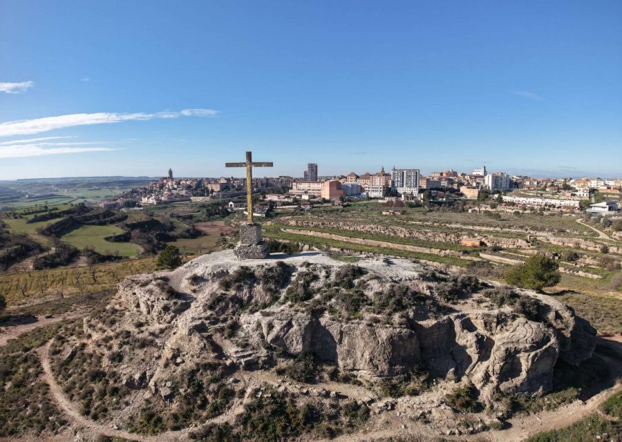 Vista aèria del turó rocós de les Forques, coronat per una gran creu de fusta, amb els camps de la Segarra a l'esquerra i el nucli de Cervera al fons