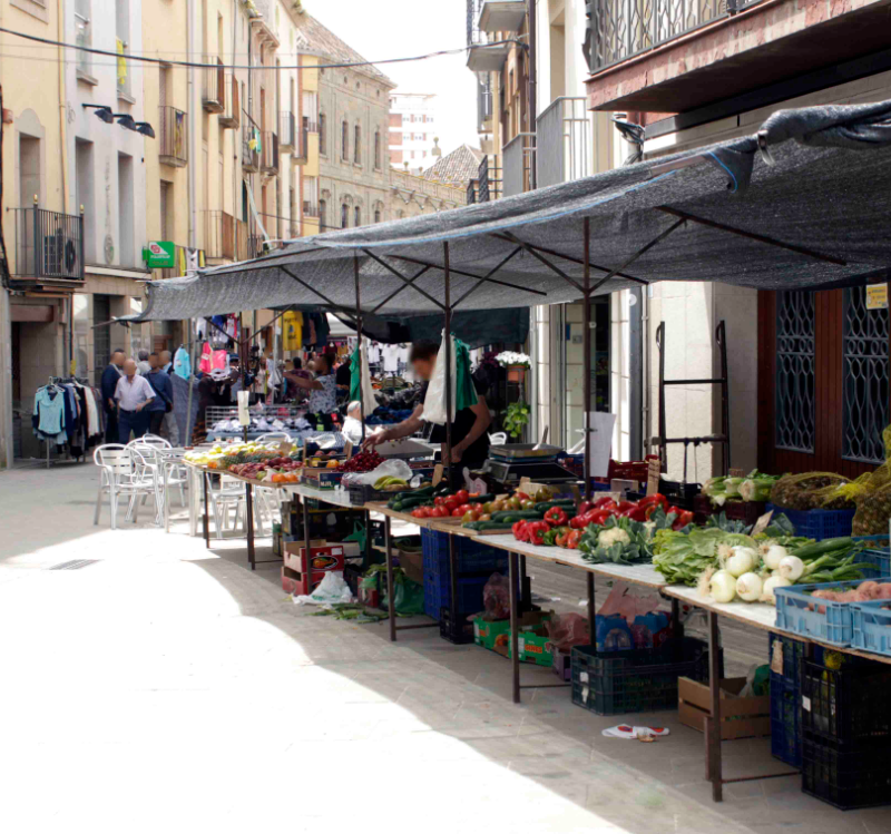 Parades del mercat setmanal de Cervera al carrer, amb caixes de verdura i fruita sobre taulells i tendals de tela fent ombra