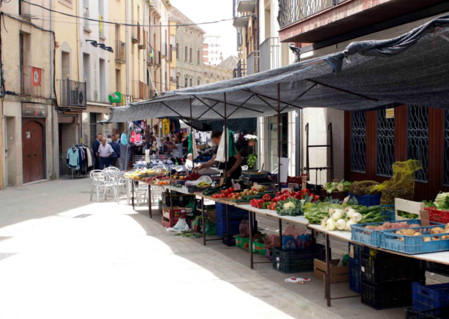 Parades del mercat setmanal de Cervera al carrer, amb caixes de verdura i fruita sobre taulells i tendals de tela fent ombra