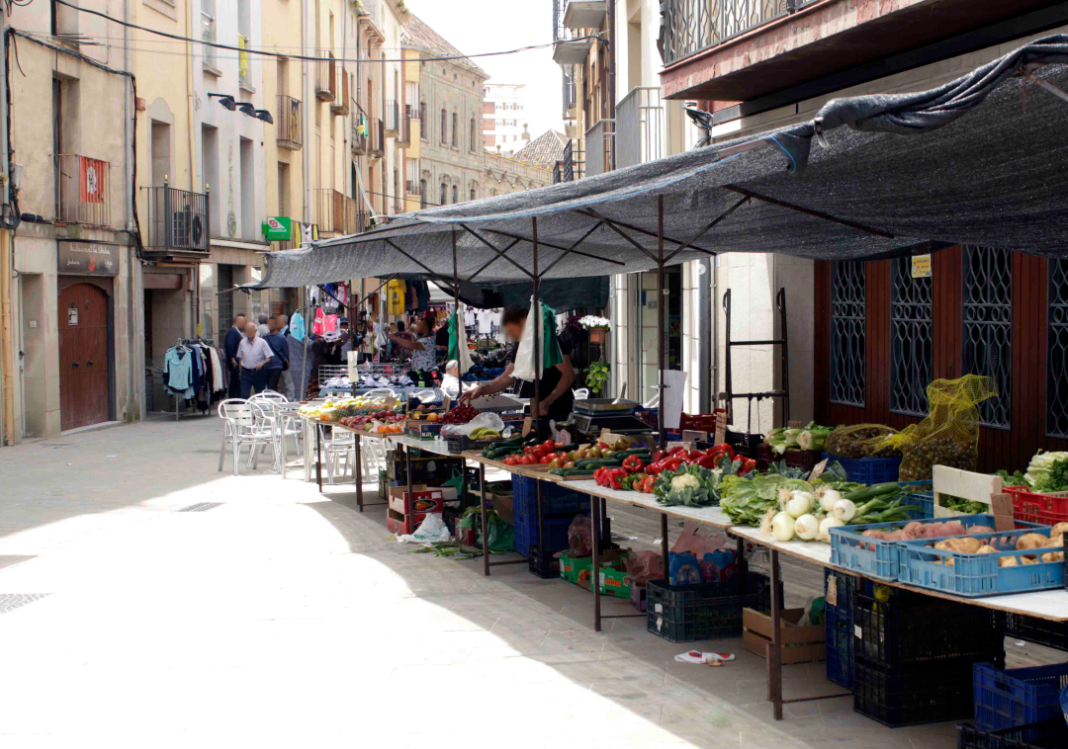 Parades del mercat setmanal de Cervera al carrer, amb caixes de verdura i fruita sobre taulells i tendals de tela fent ombra