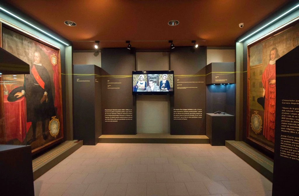 Sala d'exposició del Museu Comarcal de Cervera, amb dos grans retrats històrics emmarcats als murs laterals, plafons de text i una pantalla de vídeo al centre