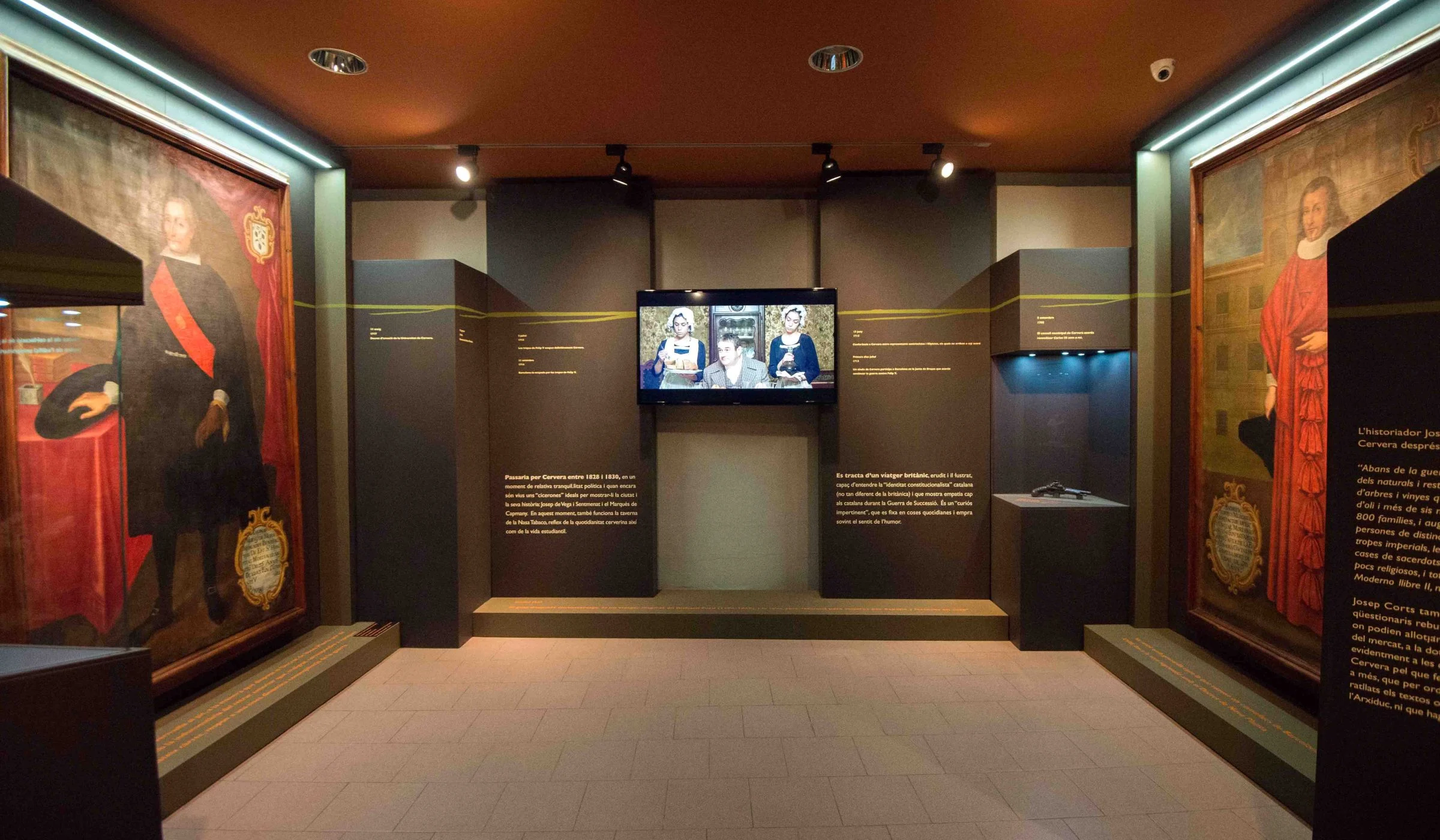 Sala d'exposició del Museu Comarcal de Cervera, amb dos grans retrats històrics emmarcats als murs laterals, plafons de text i una pantalla de vídeo al centre