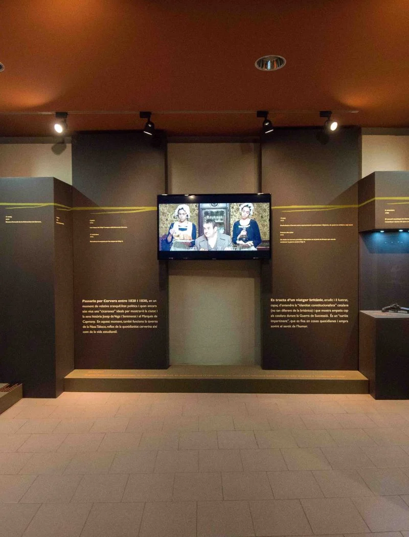 Sala d'exposició del Museu Comarcal de Cervera, amb dos grans retrats històrics emmarcats als murs laterals, plafons de text i una pantalla de vídeo al centre