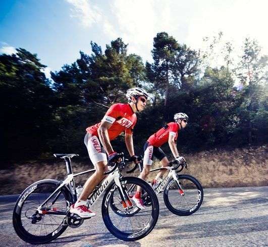 Dos ciclistes amb equipació vermella i bicicleta de carretera pedalant per una carretera asfaltada voltada de pins