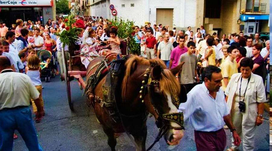 Cercavila de Sant Magí pel carrer: un cavall guarnit amb arreus estira un carro amb persones vestides de pubilla, envoltat de gent que segueix la comitiva