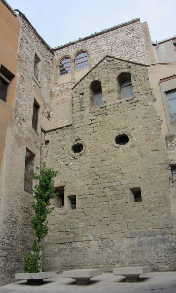 Façana de pedra de l’antiga església de Sant Joan de Jerusalem de Cervera, amb finestres romàniques i bancs a la plaça.