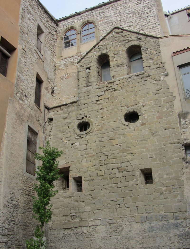 Façana de pedra de l’antiga església de Sant Joan de Jerusalem de Cervera, amb finestres romàniques i bancs a la plaça.
