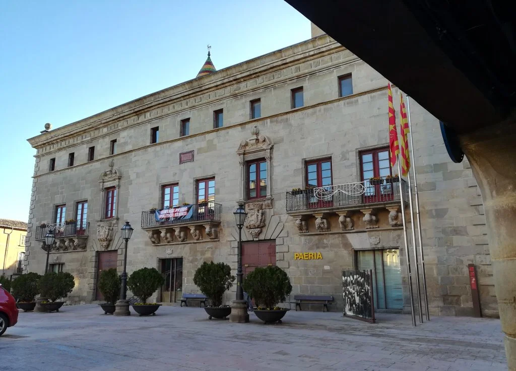 Façana de pedra de la Paeria de Cervera, amb balcons esculpits, testos a la plaça i un porxo en primer terme.
