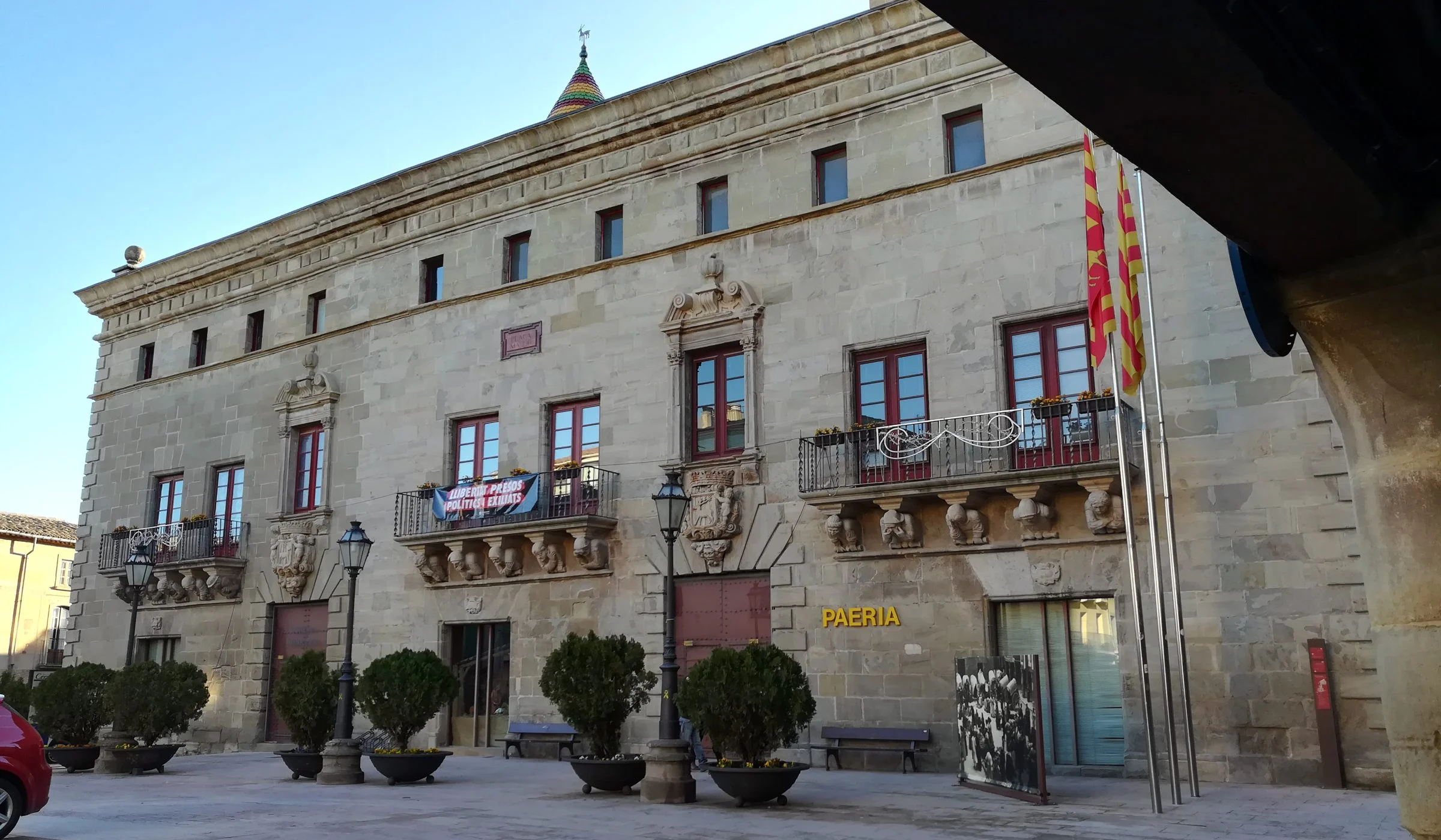 Façana de pedra de la Paeria de Cervera, amb balcons esculpits, testos a la plaça i un porxo en primer terme.