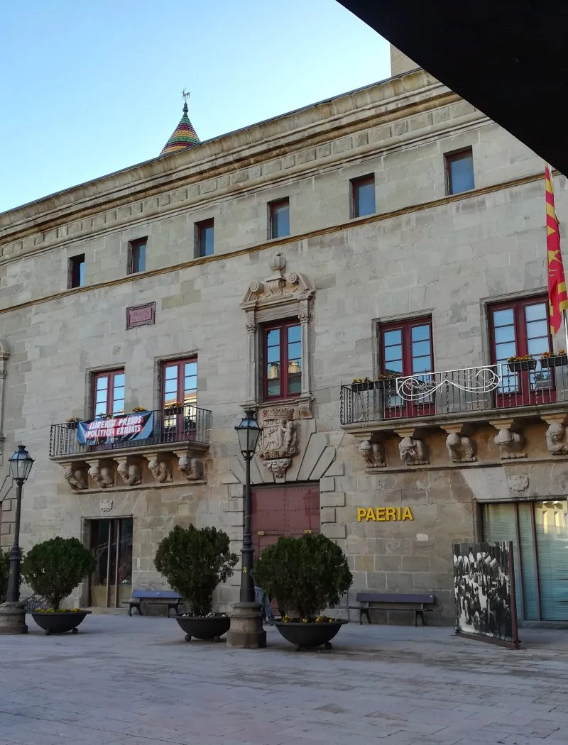 Façana de pedra de la Paeria de Cervera, amb balcons esculpits, testos a la plaça i un porxo en primer terme.